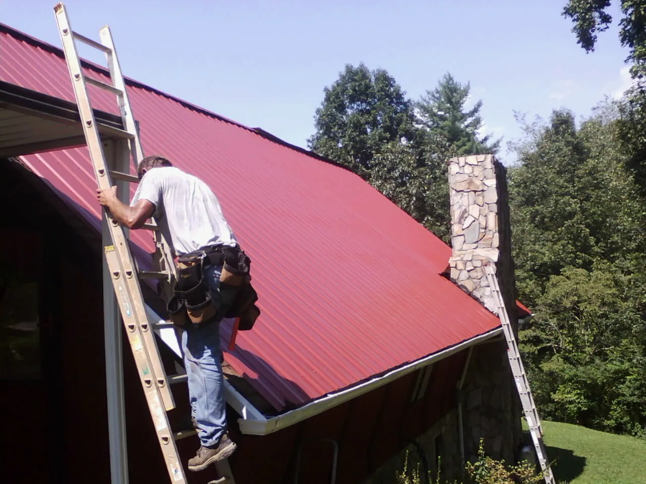 Roofer on a ladder beside a red metal roof and stone chimney — Soffit & Fascia Repair by Roofing Brothers in Clay City